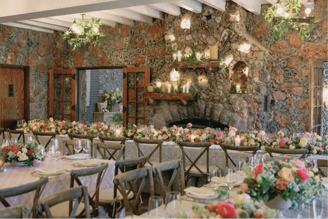 A long table dining table with floral arrangements next to a stone fireplace